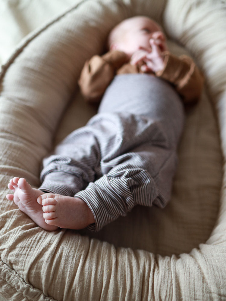 Baby in einem weichen, neutralen Baby-Nest, trägt Babyknicker Bio-Baumwolle - Schoko Ringel mit gestreiften Hosen. Der lockere Schnitt bietet Bewegungsfreiheit; elastische Bündchen sorgen für bequemen Sitz.