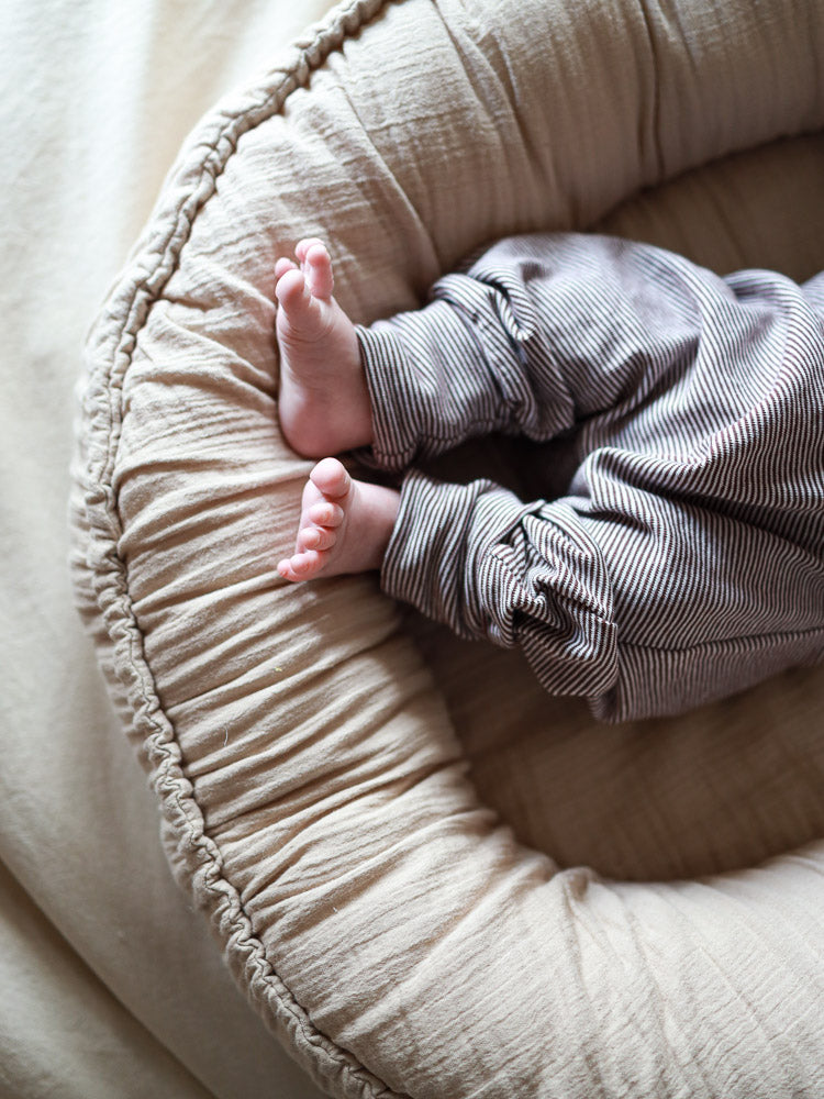 Babyknicker Bio-Baumwolle - Schoko Ringel: Nahaufnahme von Babybeinen in grauweiß gestreiften Hosen, geborgen in einem weichen, beigen Babynest.