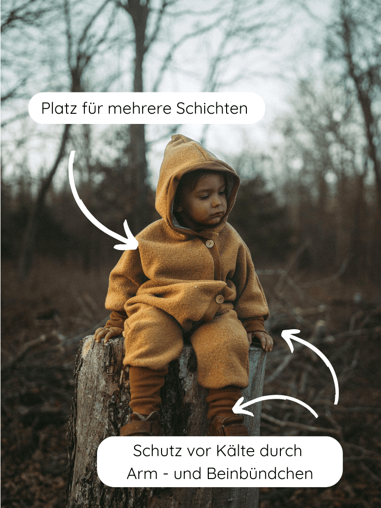 Kleinkind im warmen Kuschelanzug Wollwalk - Honig, sitzt auf Baumstumpf. Anzug mit Platz für Schichten, Arm- und Beinbündchen schützen vor Kälte. Ideal für Kita und Spaziergänge.