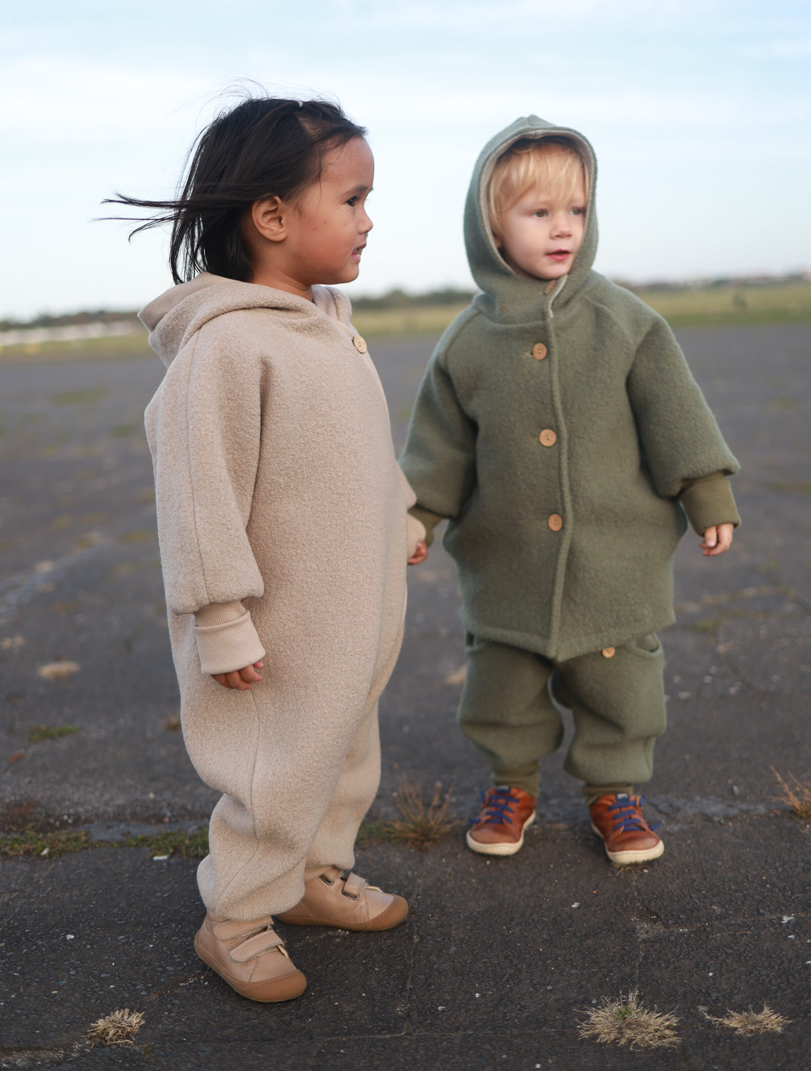 Zwei Kinder tragen den Kuschelanzug Wollwalk - Wollwalkanzug in der Farbe Hafer, ideal für Frühling und Winter mit atmungsaktivem Bio-Jersey-Futter, langen Bündchen und Wichtelkapuze.