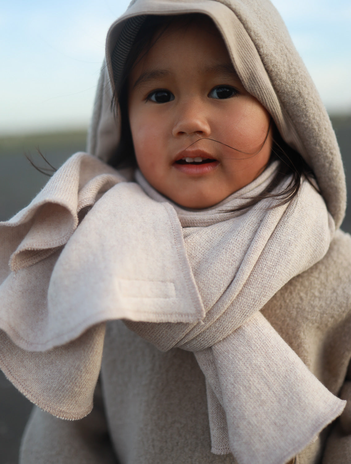 Kind trägt den Wollwalkanzug - Hafer und das Musselintuch Sand mit Kapuze und eng anliegenden Bündchen, ideal für kalte Temperaturen und Bewegungsfreiheit. Perfekt für Babytragen.