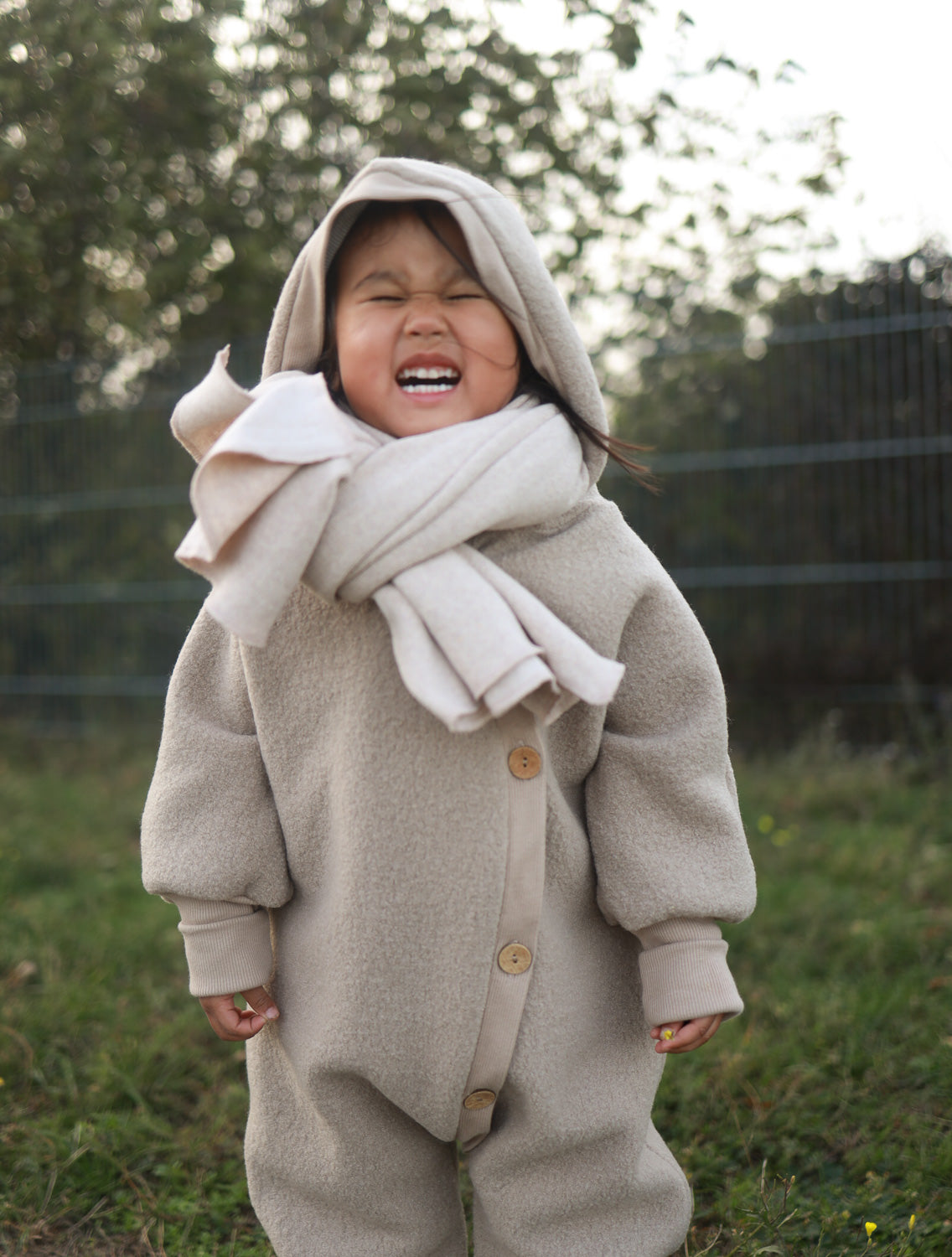 Mädchen in einem Kuschelanzug Wollwalk - Wollwalkanzug in Beige mit Kapuze und Schal, ideal für kühle Jahreszeiten, schützt vor Kälte und bietet Bewegungsfreiheit.