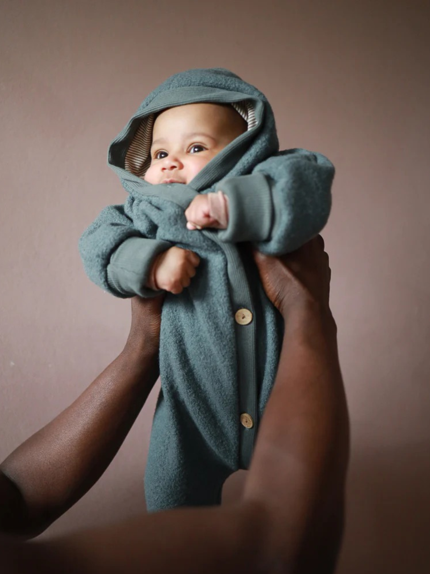 Ein Baby trägt den  Wollwalk Overall - Taube mit Kapuze und Holzknöpfen, sanft gehalten von Erwachsenen. Der Anzug bietet Wärme und Bewegungsfreiheit dank der Schurwolle und Bio-Jersey-Futter.