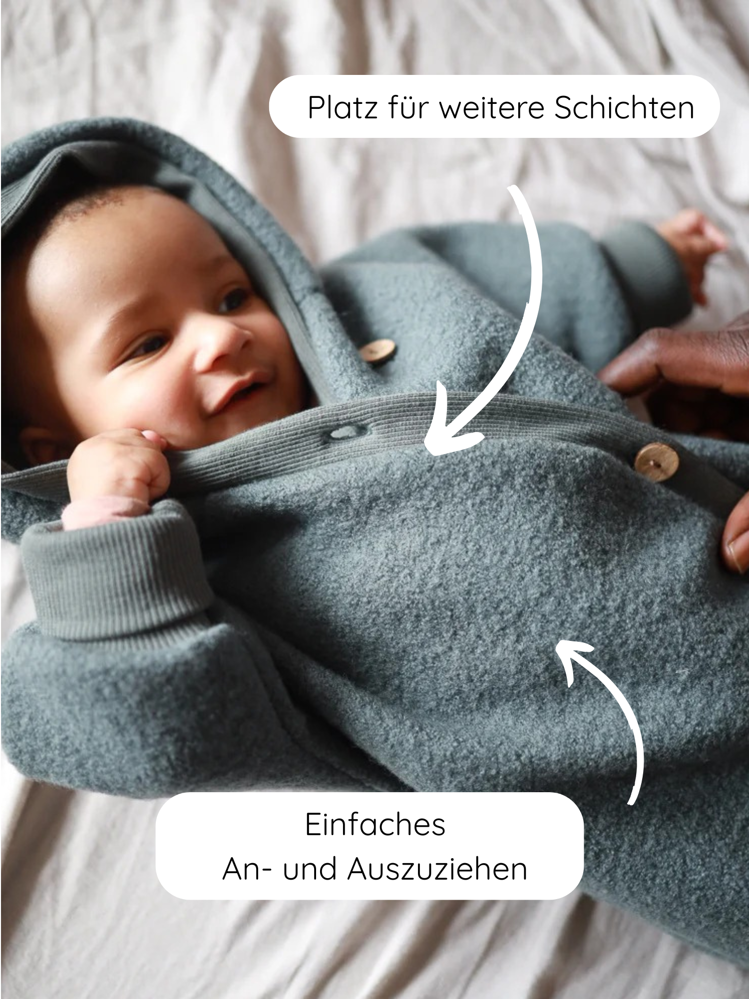 Ein lächelndes Baby liegt im Wollwalk Overall für Babys und Kinder in Taube, einem graugrünen, gefütterten Wollwalk-Einteiler mit Holzknöpfen, der Platz für Schichten bietet und einfach an- und auszuziehen ist.