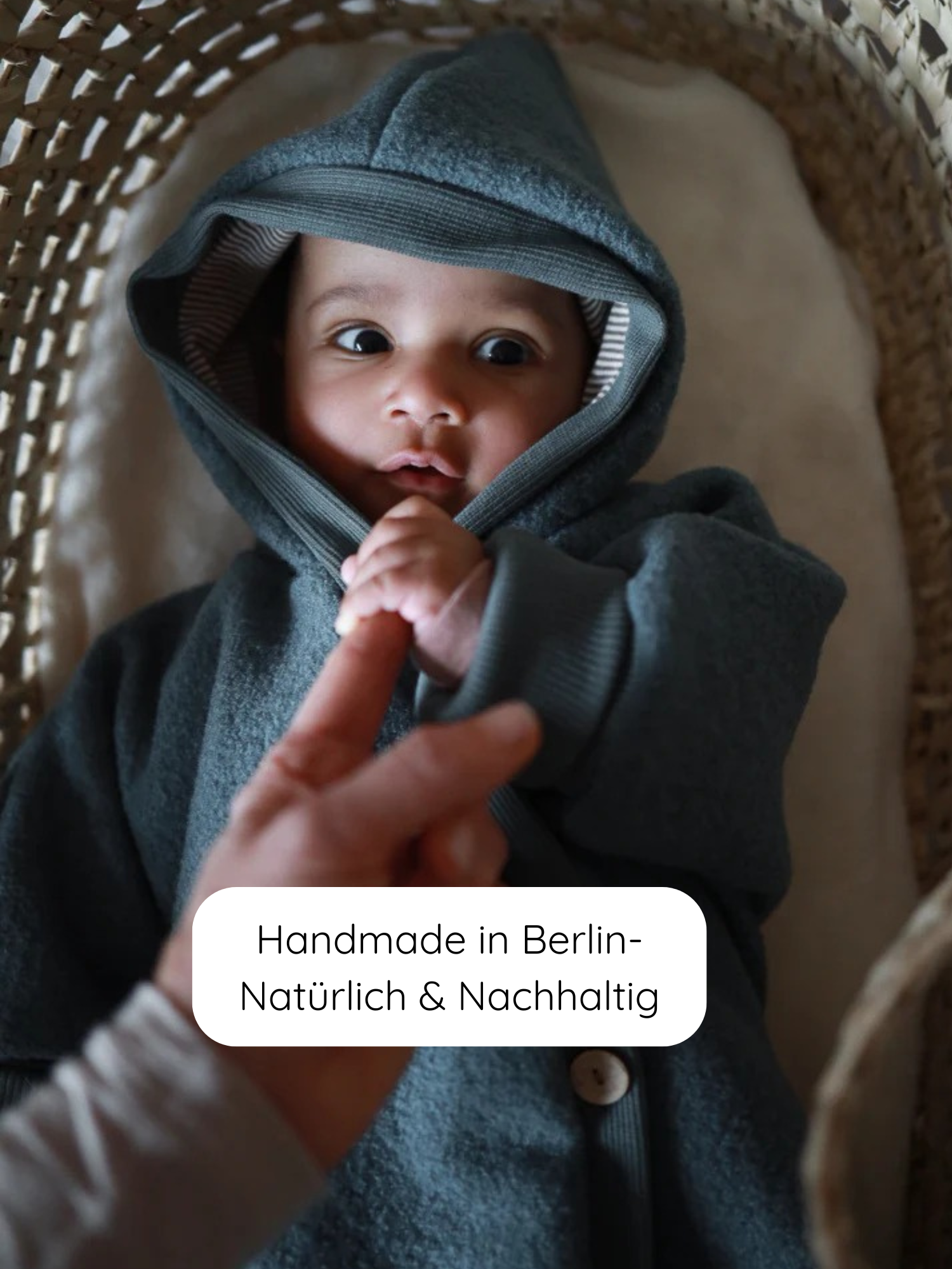 Baby in einem Kuschelanzug Wollwalk - Taube, liegt in einem Korbbett. Eine Hand berührt sanft die Hand des Babys. Label zeigt Handmade in Berlin – Natürlich & Nachhaltig.
