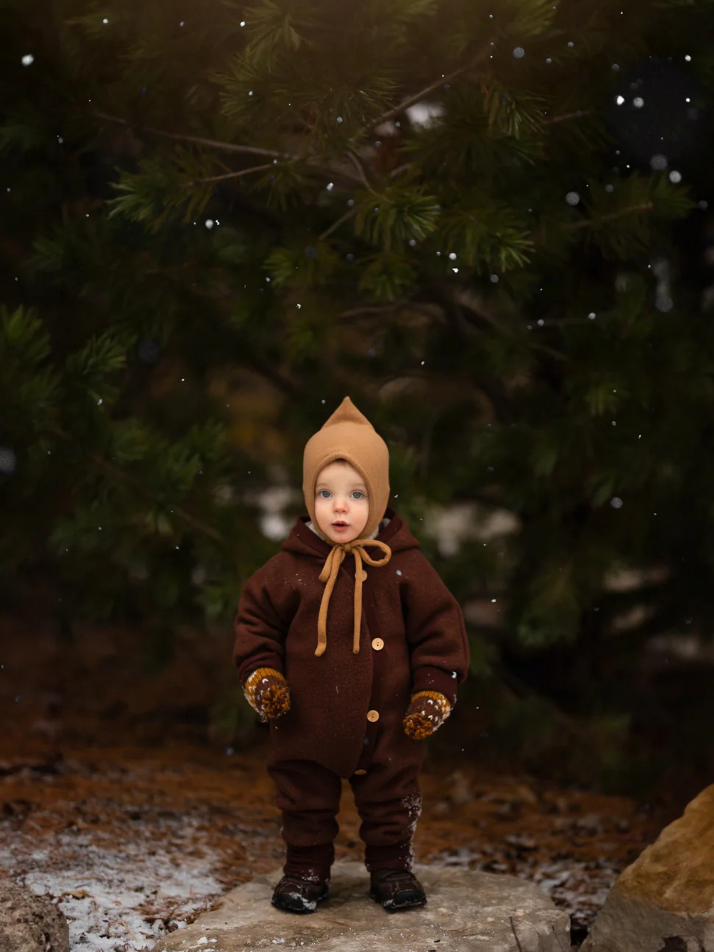 Niño pequeño con mono de lana afelpada Wollwalk color chocolate, mono marrón de una pieza con botones, manoplas a juego y gorro tipo bonnet beige, de pie sobre una piedra en un bosque invernal, rodeado de copos de nieve flotantes.