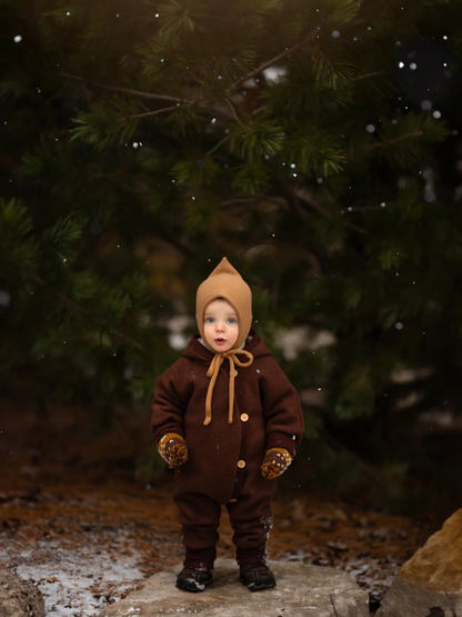 Niño pequeño con mono de lana afelpada Wollwalk color chocolate, mono marrón de una pieza con botones, manoplas a juego y gorro tipo bonnet beige, de pie sobre una piedra en un bosque invernal, rodeado de copos de nieve flotantes.