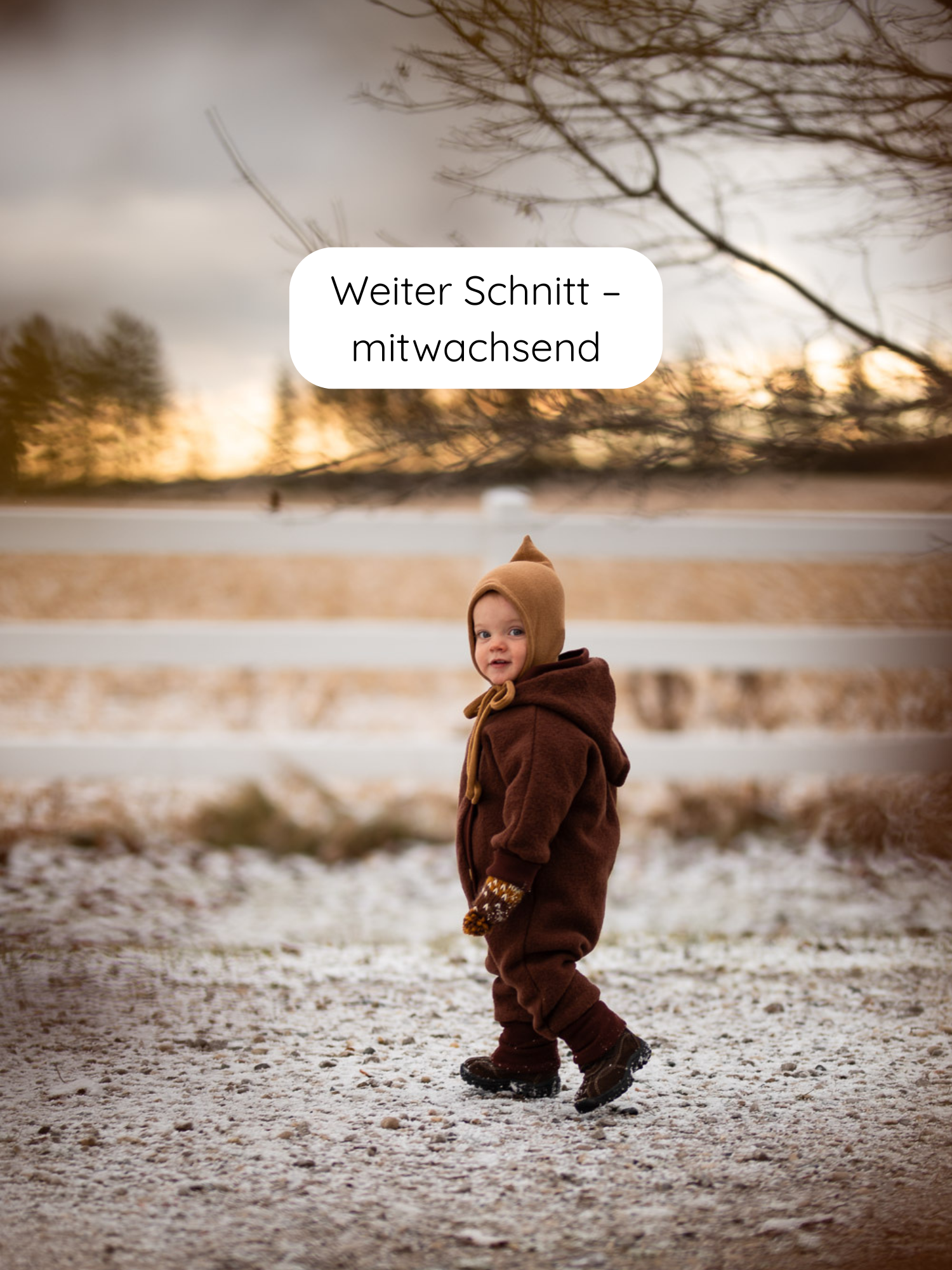 Kleinkind in einem Kuschelanzug Wollwalk - Schoko, einem warmen, schokobraunen Einteiler mit spitzer Kapuze, steht auf leicht verschneitem Boden, ideal für kalte Tage, mitwachsend durch Krempelbündchen.