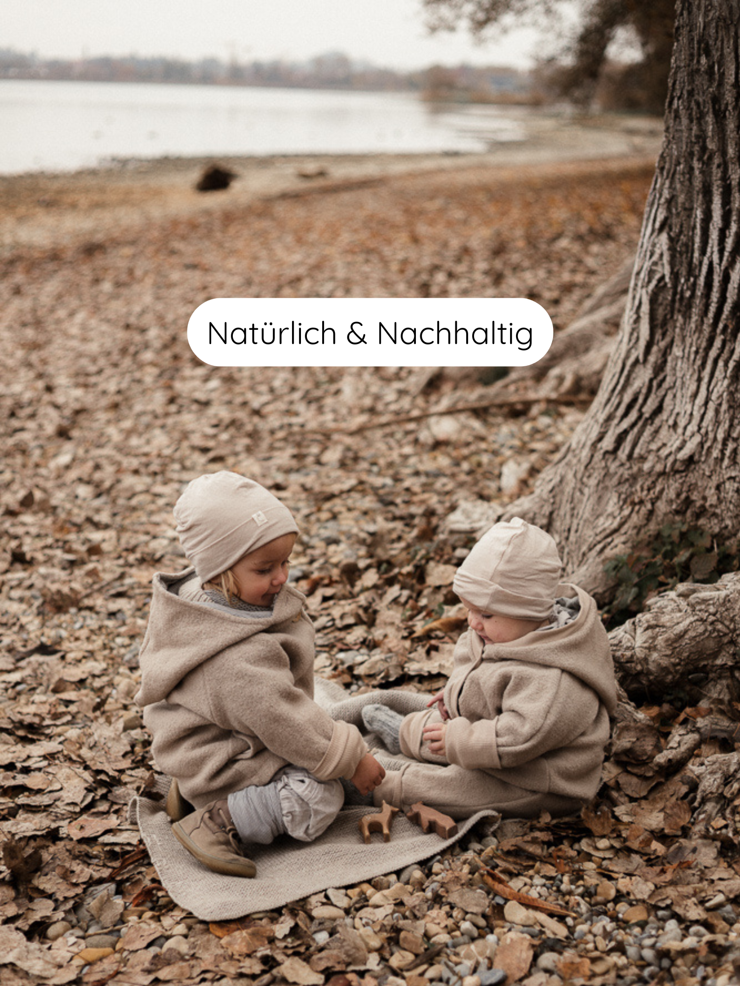 Zwei Kleinkinder im Kuschelanzug Wollwalk - Sand, sitzen auf einer Decke, spielen mit Holzfiguren, unter einem Baum. Der Anzug ist aus Schurwolle mit Bio-Jersey-Futter, natürlich und nachhaltig.