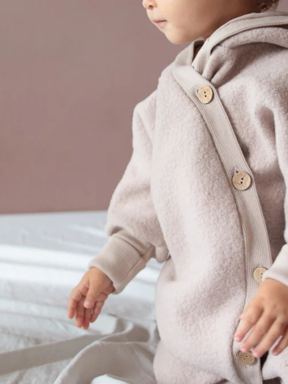 The Sand wool-walk overall shows a toddler in a beige, softly lined one-piece with wooden buttons, ideal for cool days, offering freedom of movement and protection from the cold.