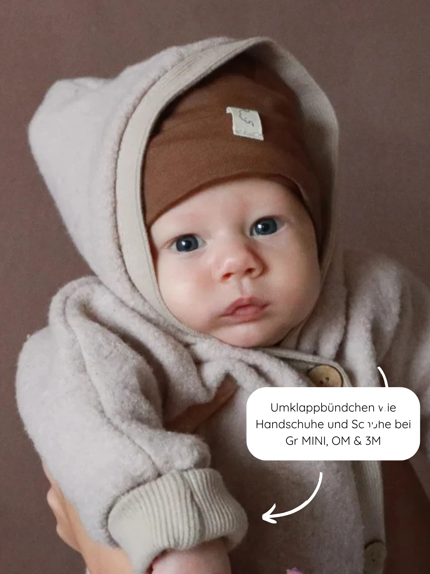 Kleinkind in einem Kuschelanzug Wollwalk - Sand, mit Kapuze und brauner Mütze. Die Ärmel zeigen die Umklappbündchen, die als Fäustlinge fungieren, ideal für kaltes Wetter.