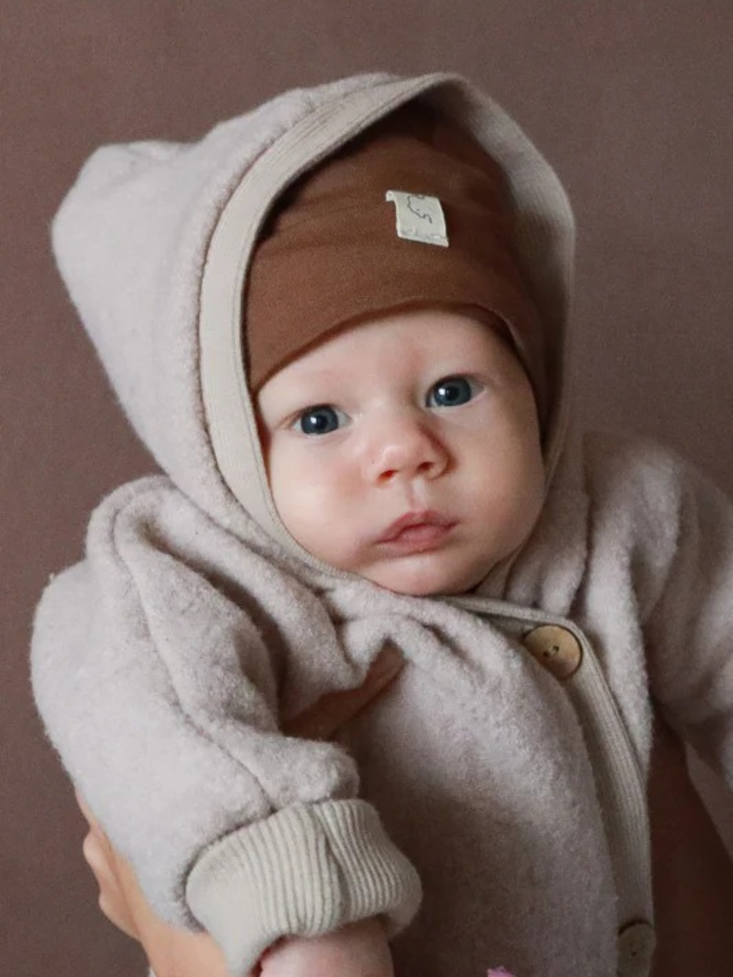 Wool Fleece Snowsuit – Sand: A baby in a soft, light beige wool fleece snowsuit with large wooden buttons and a warm hood, carried by adults. The suit provides protection and warmth while allowing freedom of movement.