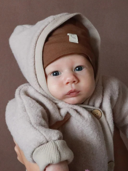 Wool Fleece Snowsuit – Sand: A baby in a soft, light beige wool fleece snowsuit with large wooden buttons and a warm hood, carried by adults. The suit provides protection and warmth while allowing freedom of movement.