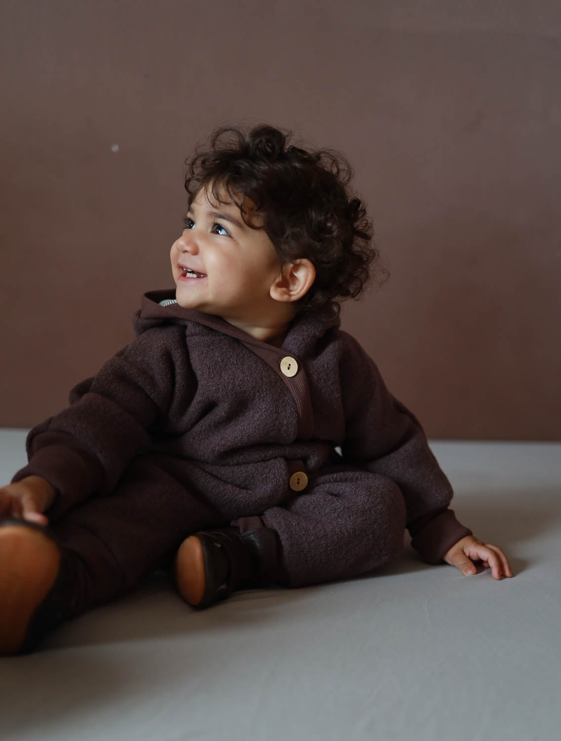Ein Baby trägt den Kuschelanzug Wollwalk - Malve, sitzt auf dem Boden. Der Anzug bietet Wärme, Bewegungsfreiheit und Schutz vor Kälte.