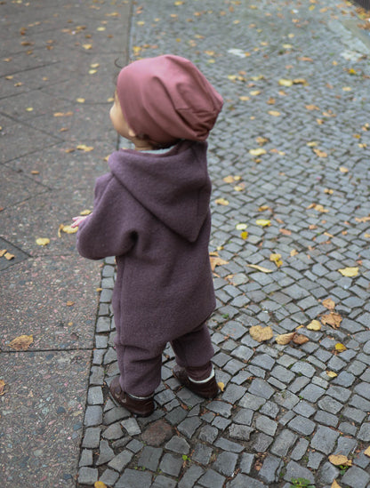 Child wearing the Wollwalk snowsuit – Mallow made from virgin wool, standing on a brick path. The overall has an elf hood, snug cuffs, and provides freedom of movement and protection from the wind. Wollwalk suit, Wollwalk overall for children, grow-with-me Wollwalk.