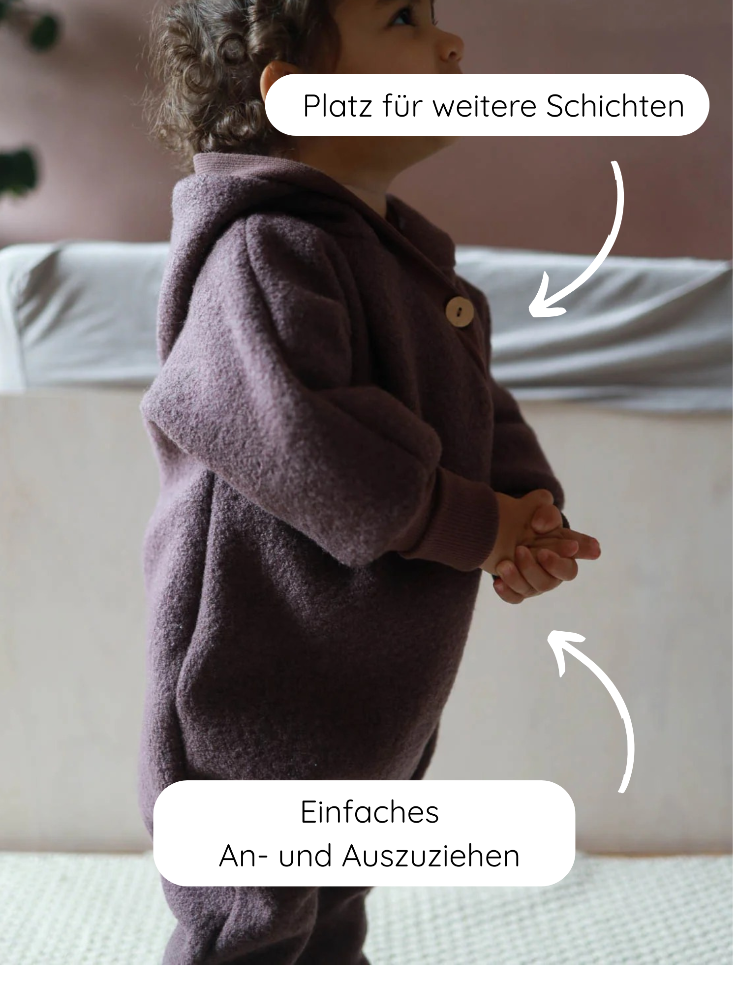 Kleinkind im Profil trägt Kuschelanzug Wollwalk - Malve mit Kapuze und Holzknopf, aus gemütlicher Schurwolle. Pfeile zeigen „Platz für weitere Schichten“ am Oberkörper und „Einfaches An- und Ausziehen“ am Bund.