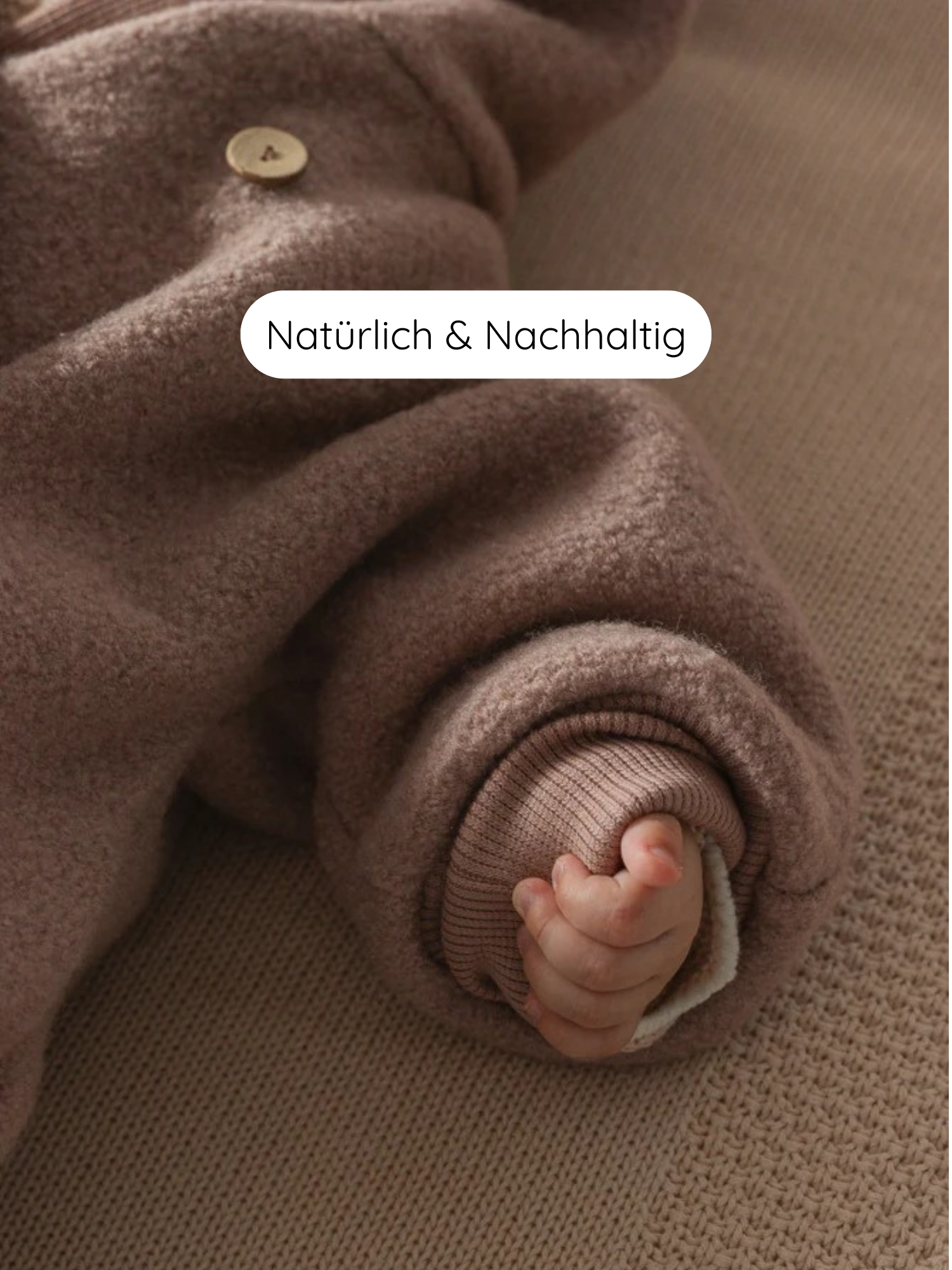 Winter Wollwalk Overall mit Teddyfutter- Puder: Nahaufnahme einer Babyhand im braunen Wollwalk-Einteiler mit Holzkopf, auf brauner Strickfläche liegend. Etikett: „Natürlich & Nachhaltig“ im Bild sichtbar.