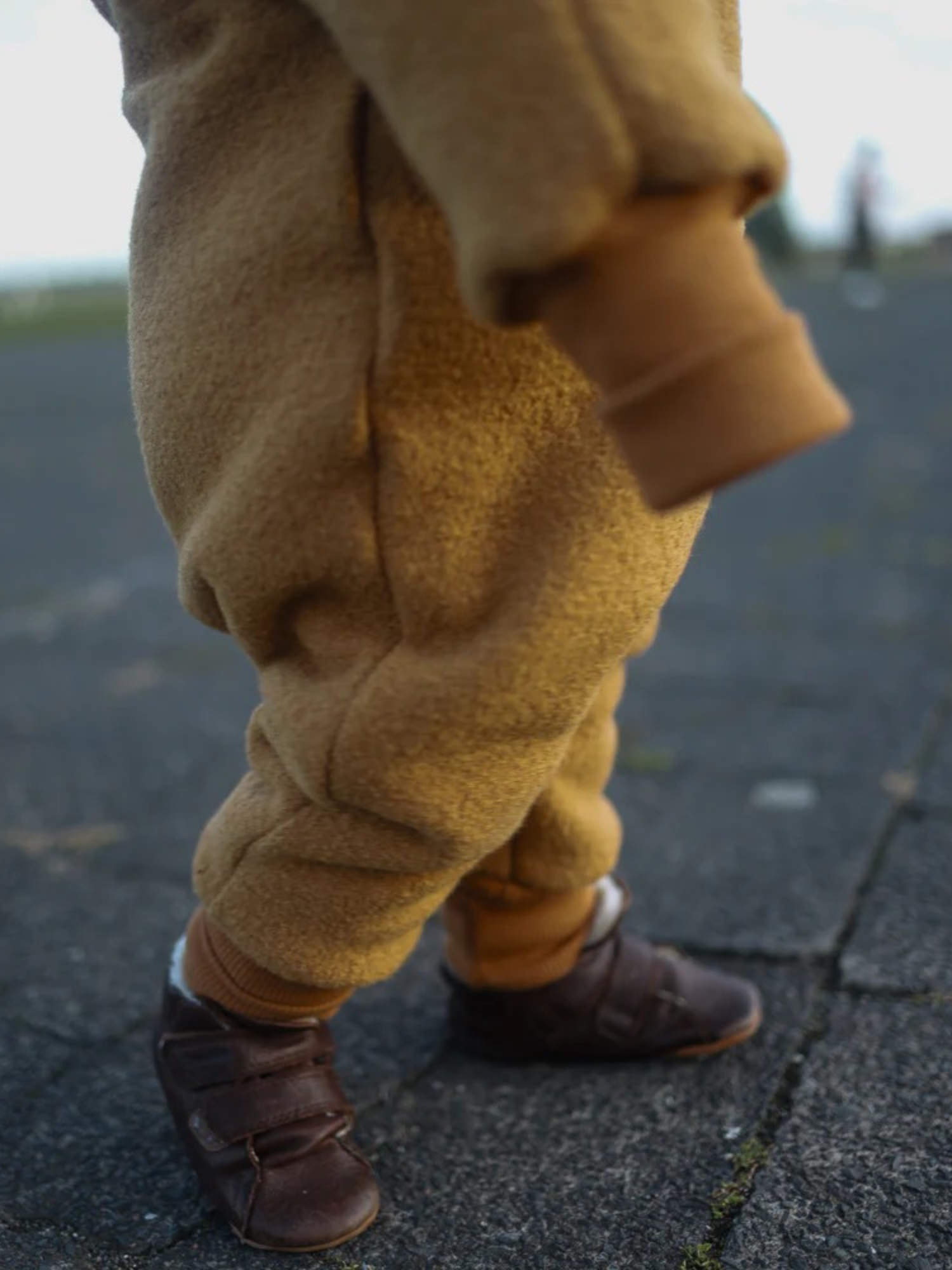 Winter Wollwalk Overall mit Teddyfutter - Honig: Nahaufnahme der unteren Körperhälfte eines Kindes in einem flauschigen, camel-braunen Overall, kombiniert mit dunkelbraunen Lederschuhen, auf einer gepflasterten Fläche stehend.