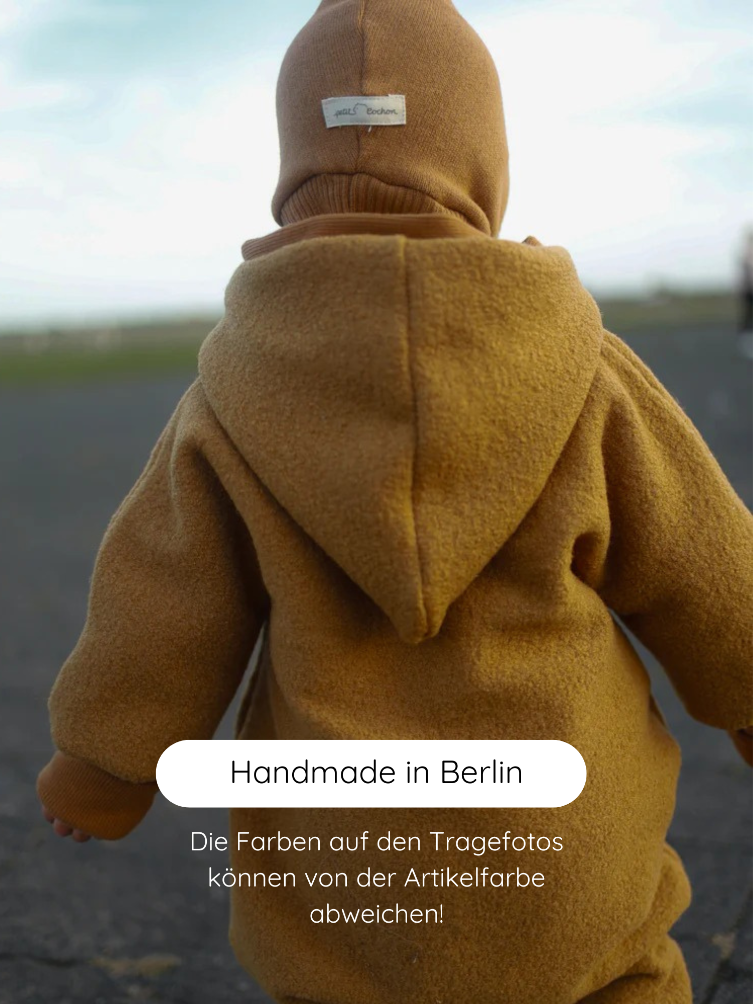 Kleinkind in einem mustardfarbenen Winter Wollwalk Overall mit Teddyfutter - Honig, zeigt die Rückseite. Der Overall hat eine spitze Kapuze und eine passende Strickmütze mit petit cochon-Etikett.