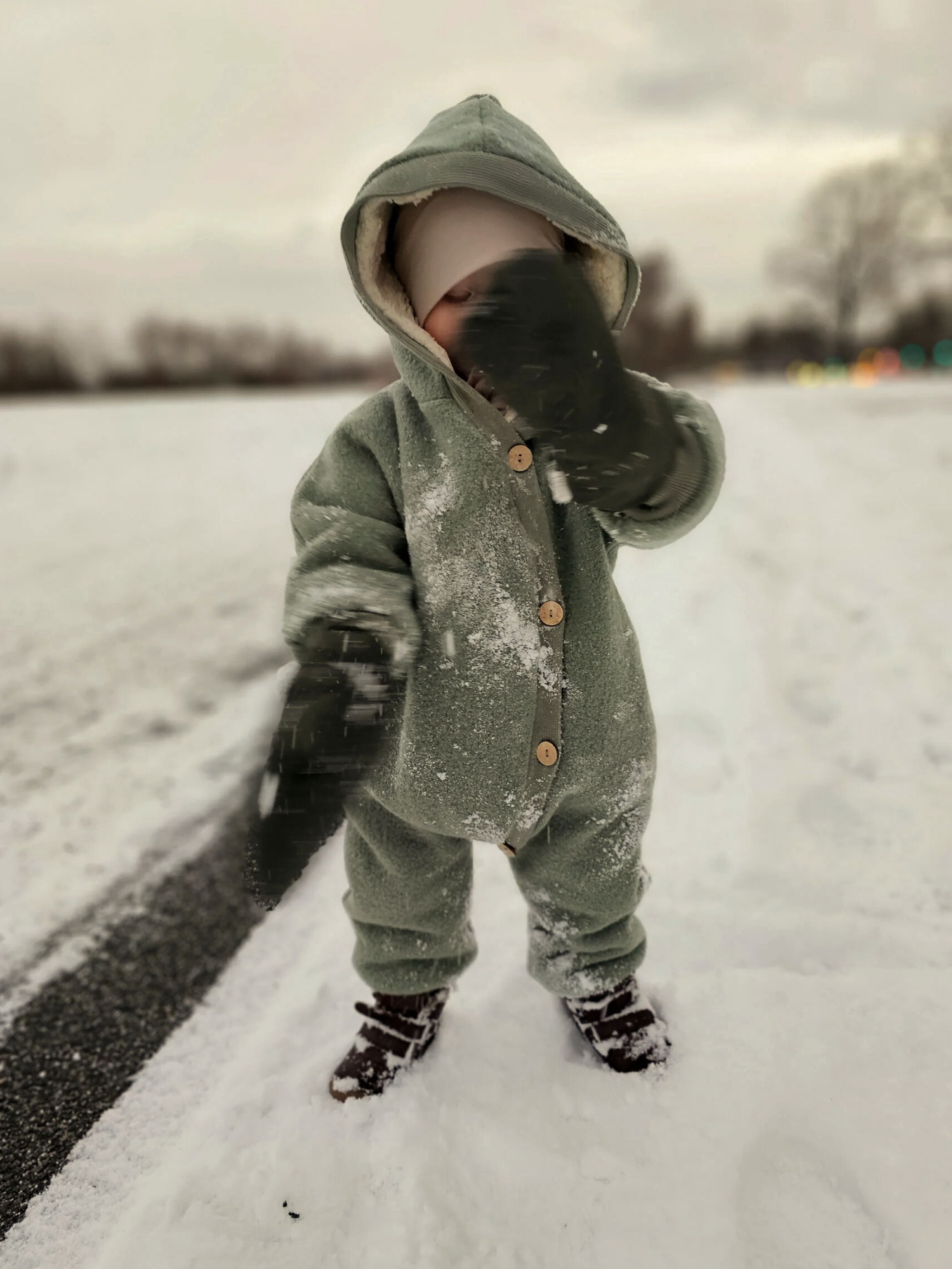 Kleinkind im Winter Wollwalk Overall mit Teddyfutter - Pistazie, trägt dunkle Stiefel und Handschuhe im Schnee, hebt eine behandschuhte Hand, um Schneeflocken abzuwischen.