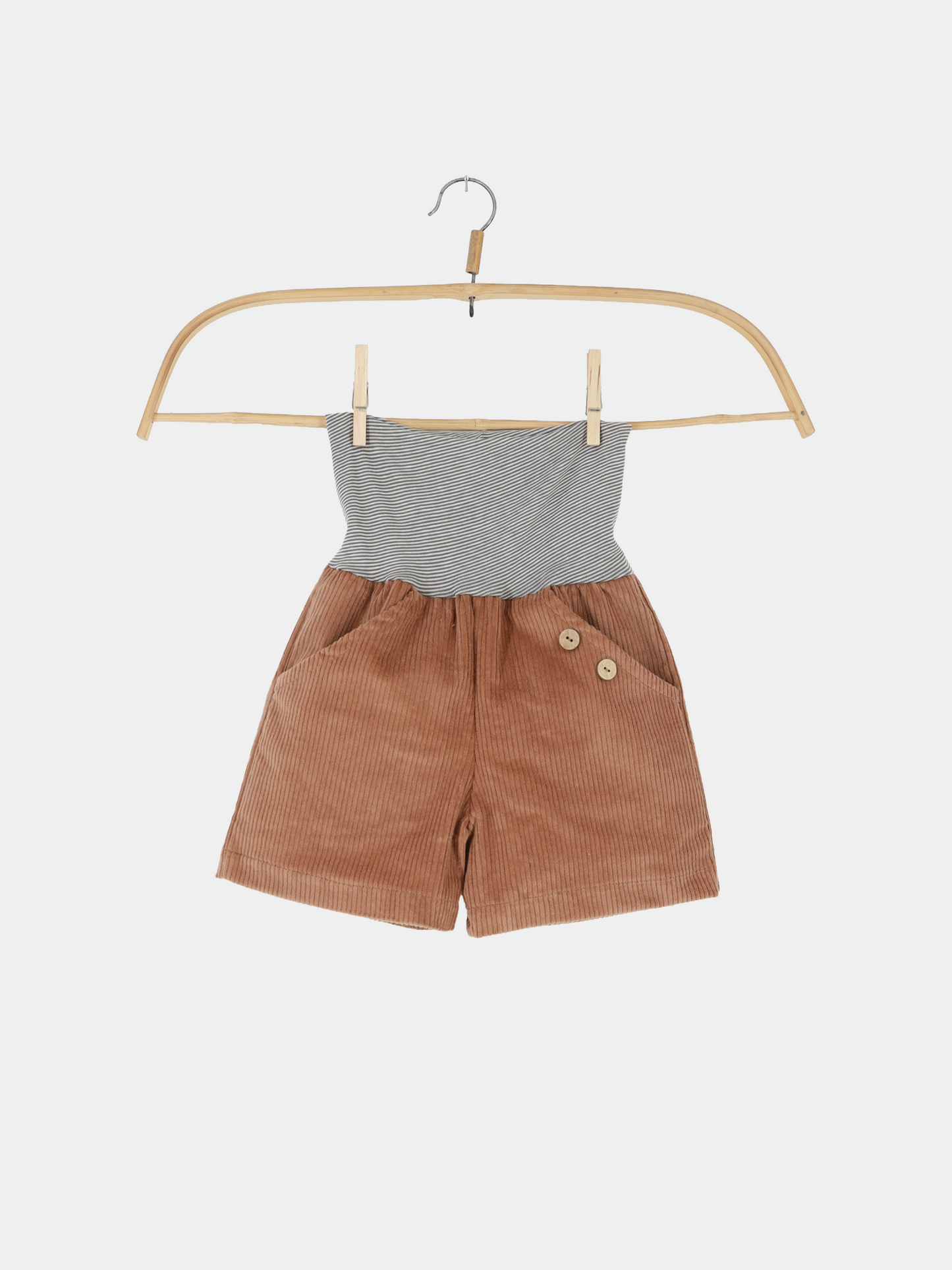 Kurze Buxe Breitcord - Weizen: Kinder-Shorts aus weichem, rostbraunem Cord mit seitlichen Taschen, zwei Holzknöpfen und einem grauweiß gestreiften, breiten Strickbund auf einem Holzkleiderbügel.