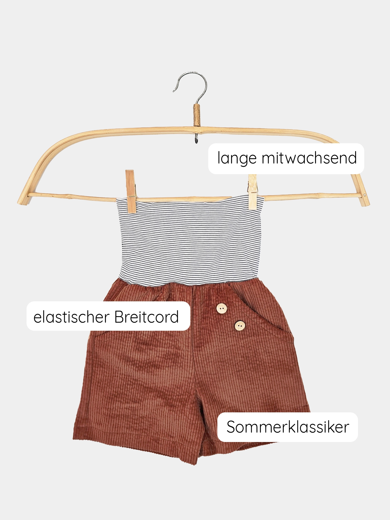 Kurze Buxe Breitcord Rost auf einem Kleiderbügel, bestehend aus braunen Knöpfen und einem gestreiften Ringelbündchen, präsentiert auf einem Holzrahmen. Nachhaltig in Berlin gefertigt aus robustem Bubblecord.