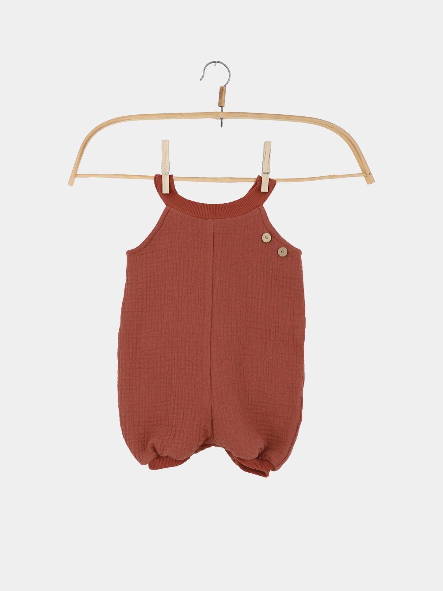 Kurze Schlupflatzhose Bio-Musselin - Terrakotta: Handgenähter Kinderromper aus strukturiertem Stoff mit zwei Holzknöpfen an der Schulter, präsentiert auf einem Holzkleiderbügel, ideal für bequeme Sommerabenteuer.