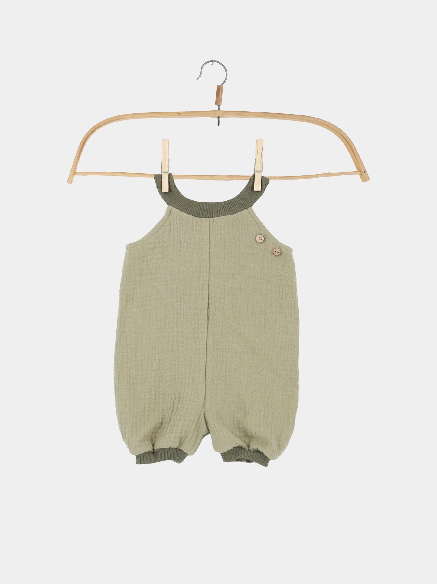 Kurze Schlupflatzhose Bio-Musselin - Salbei in crinkle Muselin-Stoff mit geripptem Besatz und zwei Holzknöpfen an der Schulter, hängend an einem Holzkleiderbügel. Handgenäht, ideal für bequeme Sommerabenteuer.