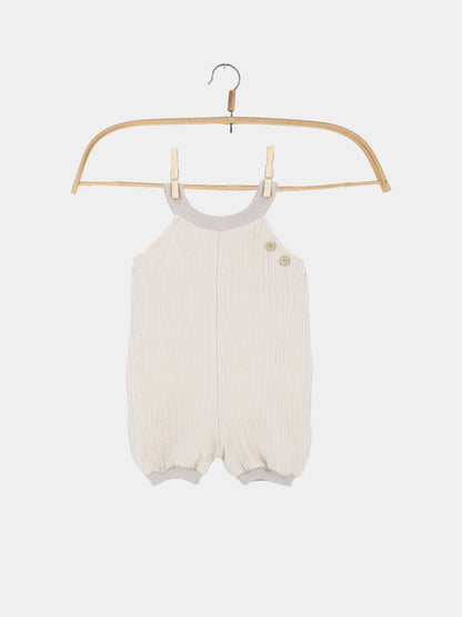 Kurze Schlupflatzhose Bio-Musselin - Sand: Ein cremefarbener, ärmelloser Baby-Strampler aus weichem Musselin mit geripptem Ausschnitt, Beinbündchen und zwei kleinen Holzknöpfen auf einer Schulter. Ideal für den Sommer.