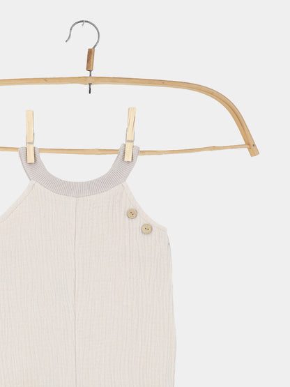 Kurze Schlupflatzhose Musselin - Sand: Ein ärmelloser Baby-Strampler aus strukturiertem Bio-Musselin, aufgehängt an einem Holzkleiderbügel mit kleinen Holzknöpfen an der Schulter, ideal für sommerliche Bewegungsfreiheit.