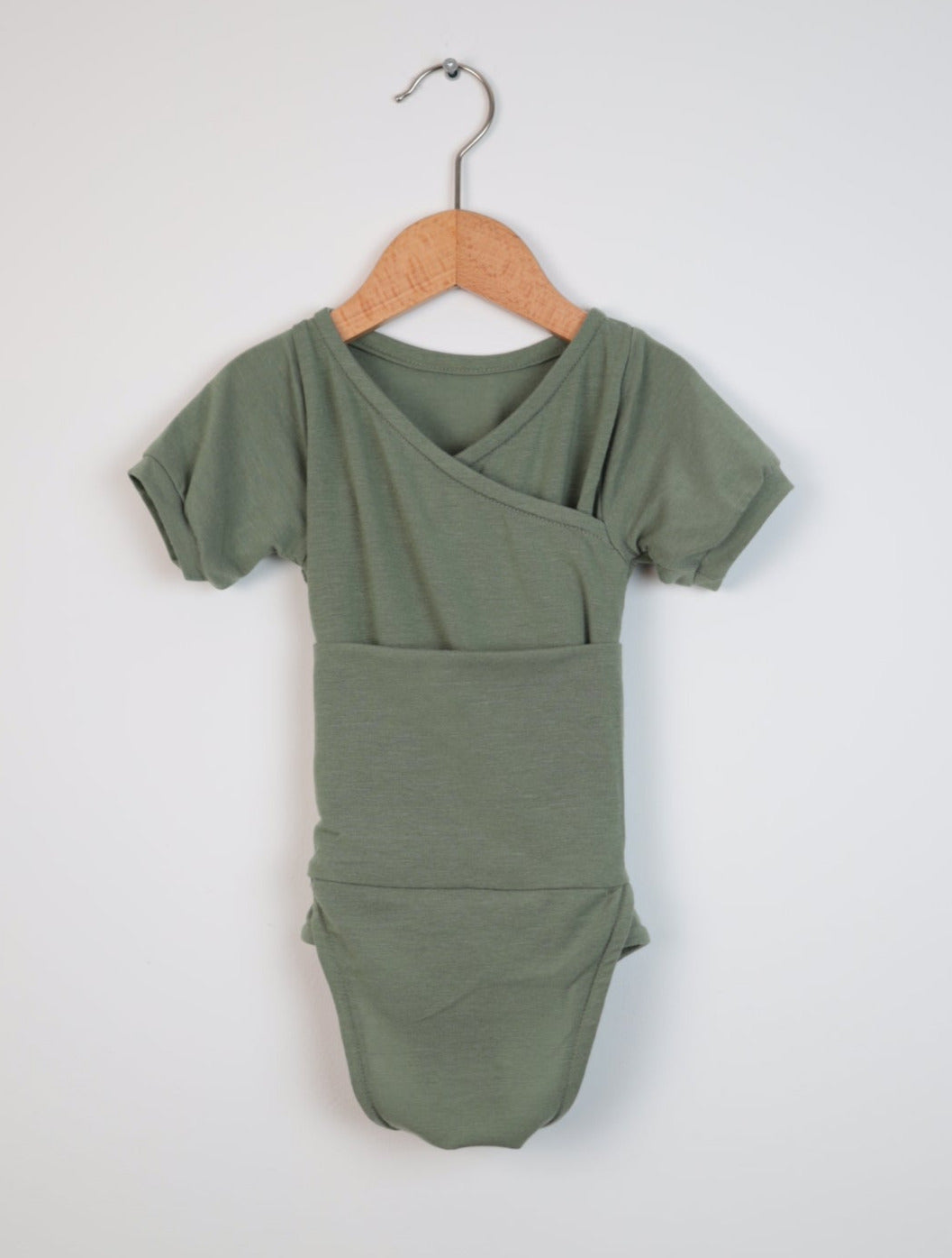 Kurzarm Loopbody Kaschmir Eukalyptus Gr. 0M, ein kimono-artig überlappendes, kurzärmeliges Baby-Body, hängt an einem Holzkleiderbügel vor einem schlichten Hintergrund. Zustand: neu.