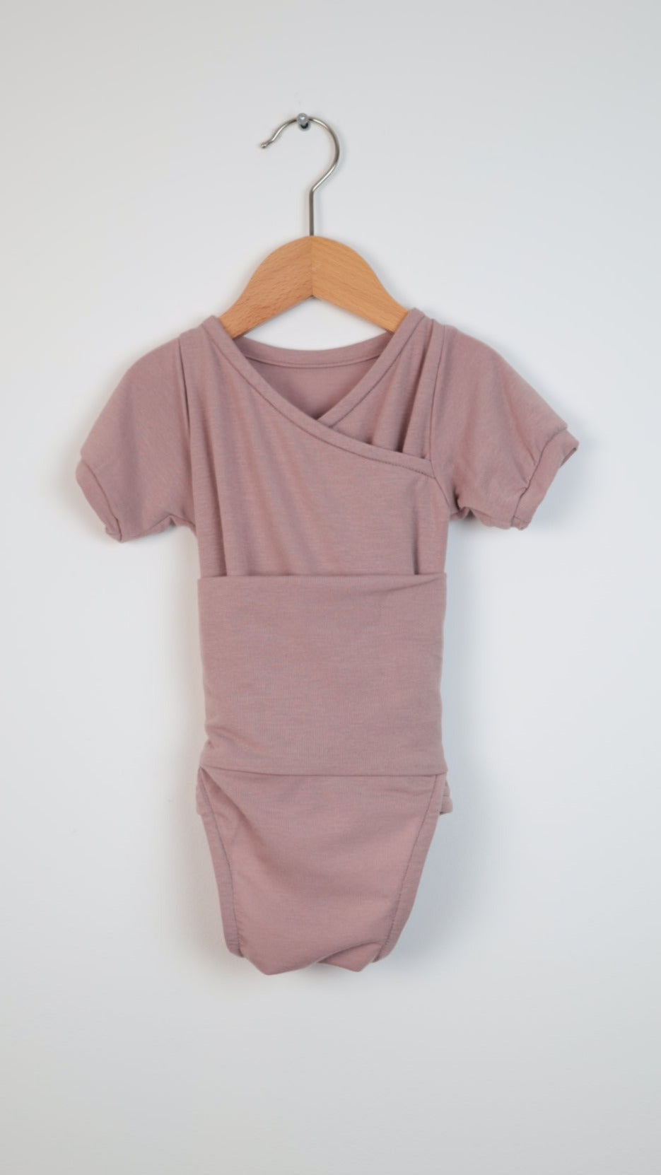 Kurzarm Loopbody Kaschmir Rosé Gr. 0M, ein Wickelbody für Neugeborene mit kurzen Ärmeln, hängt an einem Holzkleiderbügel vor einem weißen Hintergrund und präsentiert sich in einem eleganten Design.