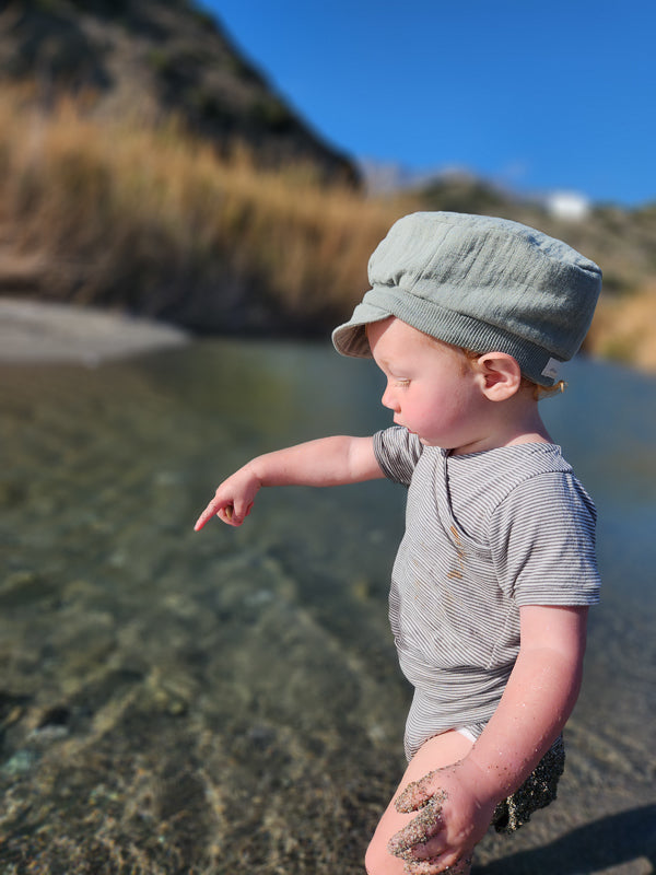 Ein Kleinkind trägt den Loopbody Kurzarm Bio-Baumwolle - Ringel Maus im Streifenmuster, steht am Wasserrand und zeigt ins Wasser, umgeben von sandigem Boden und goldener Landschaft.