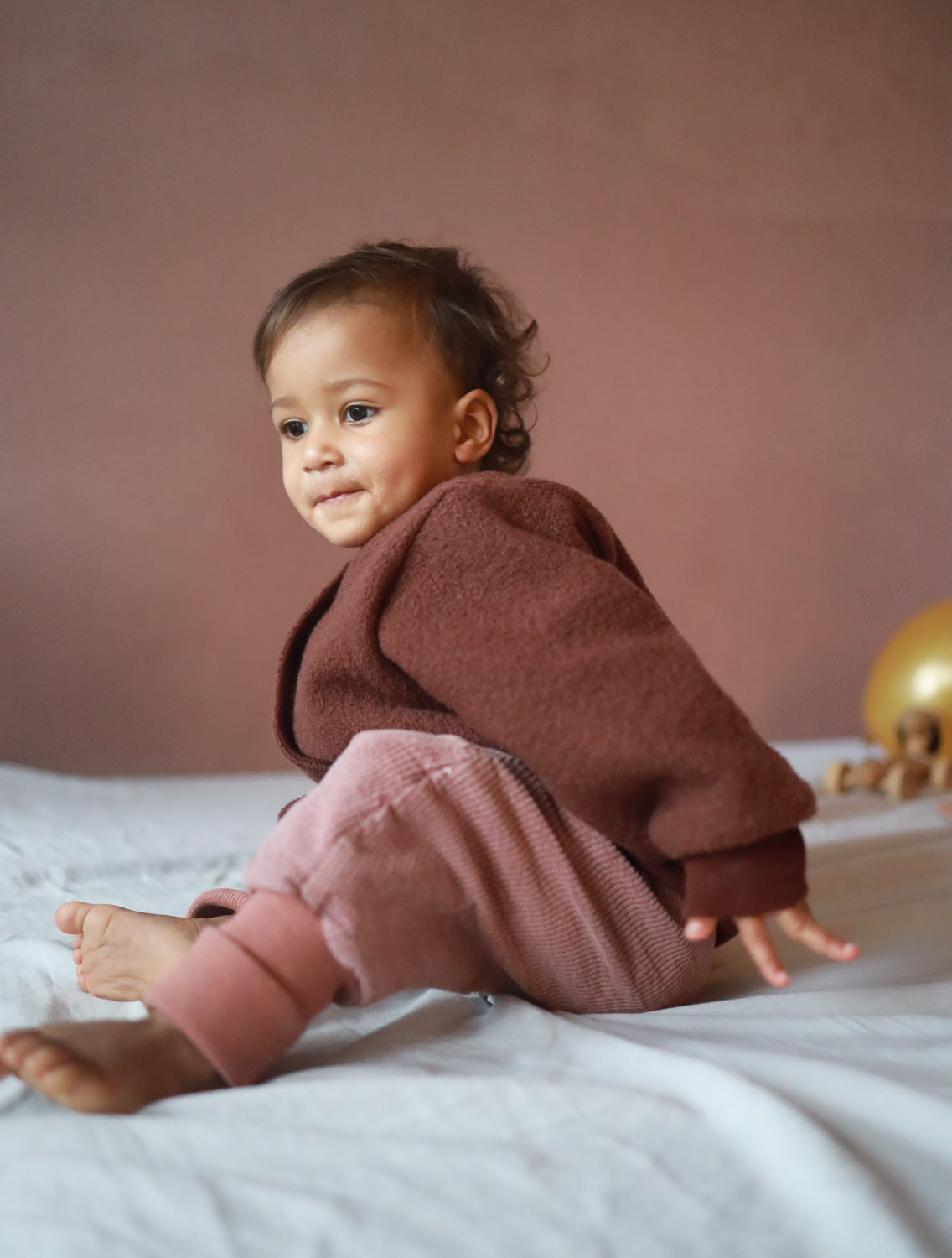Kleinkind in Knickerbocker Breitcord Stretch - Rosenholz auf Bett, trägt braune Fleecejacke, rosa Hose. Perfekt für aktive Kinder, bietet Bewegungsfreiheit und Komfort. Ideal für Übergangszeit und kühlere Tage.