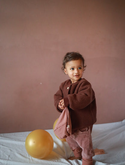 Kleinkind in Knickerbocker Breitcord Stretch - Rosenholz, steht barfuß auf einem weißen Tuch, mit weichem Stoff in der Hand, zwei goldene Ballons daneben.