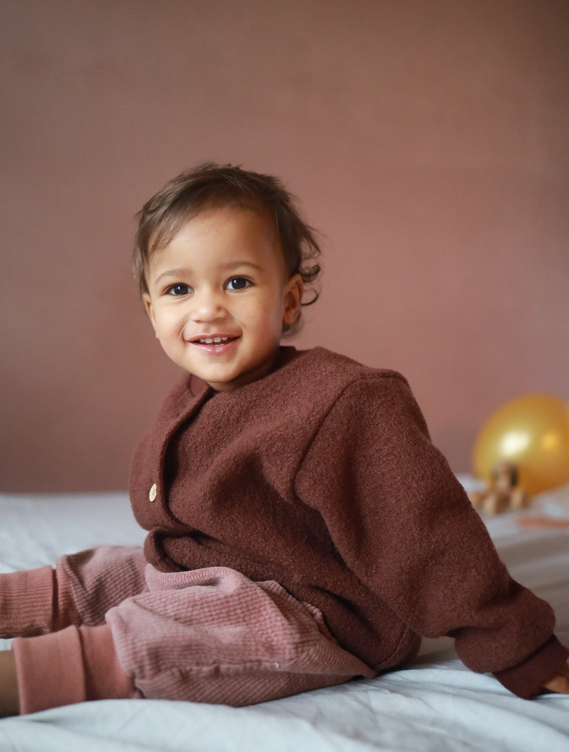 Kleinkind sitzt lächelnd mit Knickerbocker Breitcord Stretch - Rosenholz auf einem Bett, trägt einen braunen Pullover. Die weiche Hose bietet Bewegungsfreiheit und ist ideal für kühlere Tage.