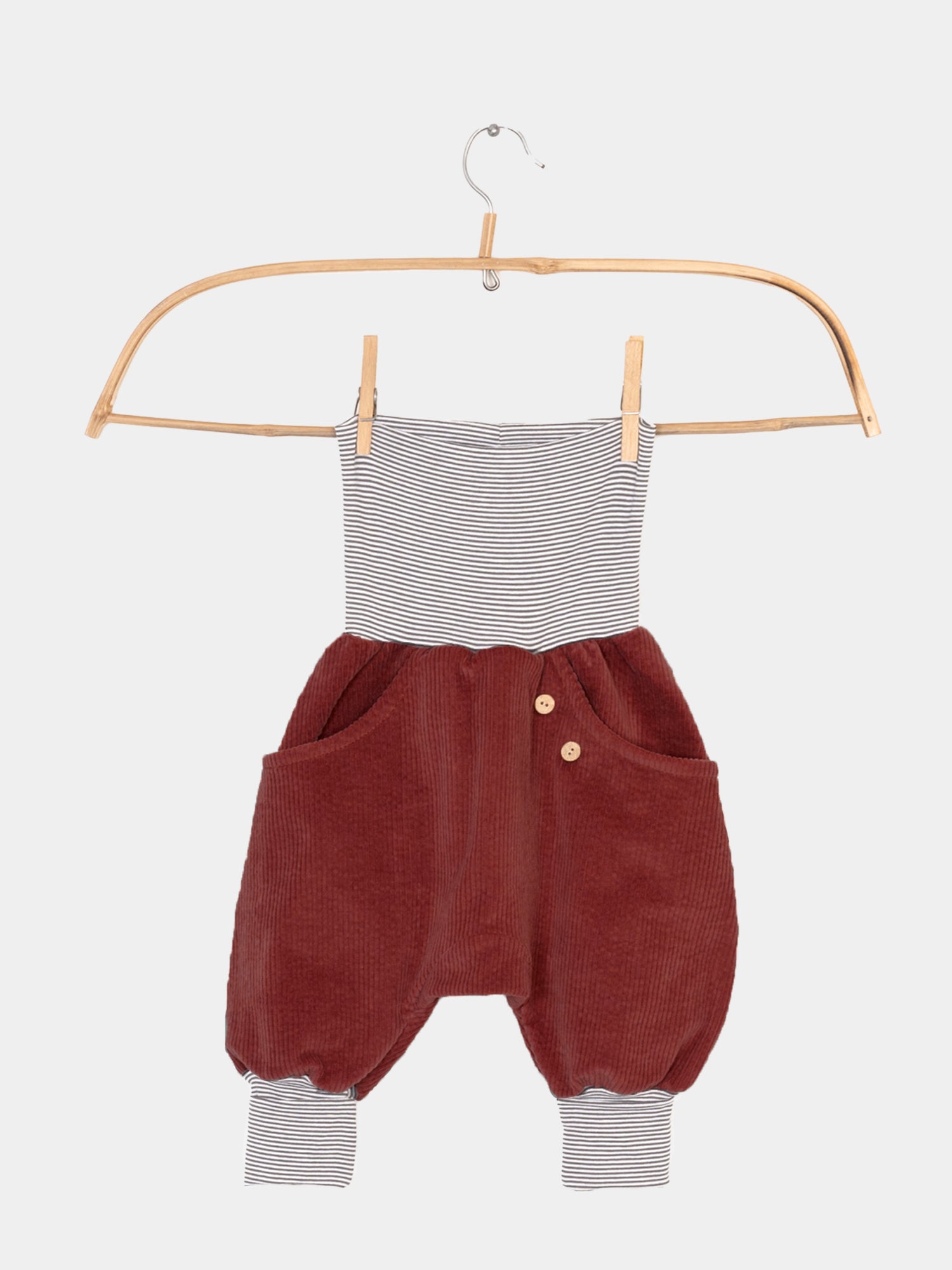 Knickerbocker Breitcord mit Ringelbündchen - Erdbeere: Kinder-Einteiler mit schwarz-weißen Streifen oben, bordeauxfarbenem Cord unten, Holzknöpfen an der Taille und gestreiften Bündchen an den Knöcheln, auf einem Holzbügel hängend.