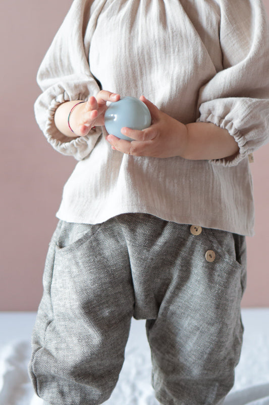Kleinkind trägt Knickerbocker Fischgrat aus Leinen/Baumwolle - mitwachsende Kinderhose - Praline, kombiniert mit cremefarbenem Oberteil, hält blaues Orb in Händen.
