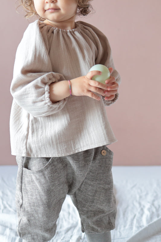 Ein Kleinkind trägt die Knickerbocker Fischgrat aus Leinen/Baumwolle - mitwachsende Kinderhose - Praline, mit grauem Fischgrätmuster, Holzknopf und elastischem Bund, hält einen grünen Ball.