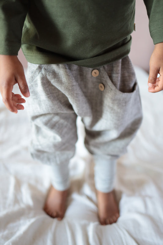 Das Bild zeigt die Knickerbocker Fischgrat aus Leinen/Baumwolle - mitwachsende Kinderhose - Praline, getragen von einem Kind. Die Hose hat einen lockeren Schnitt mit zwei sichtbaren Holzknöpfen am Bund.