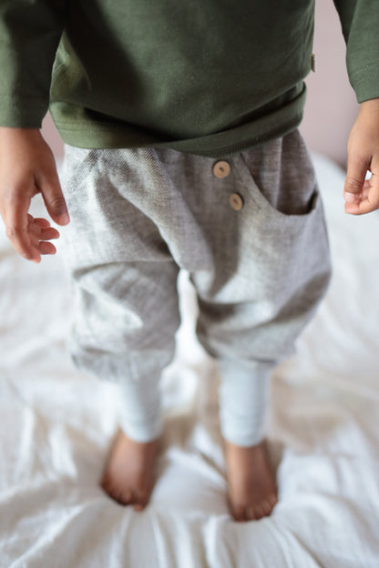 Das Bild zeigt die Knickerbocker Fischgrat aus Leinen/Baumwolle - mitwachsende Kinderhose - Praline, getragen von einem Kind. Die Hose hat einen lockeren Schnitt mit zwei sichtbaren Holzknöpfen am Bund.