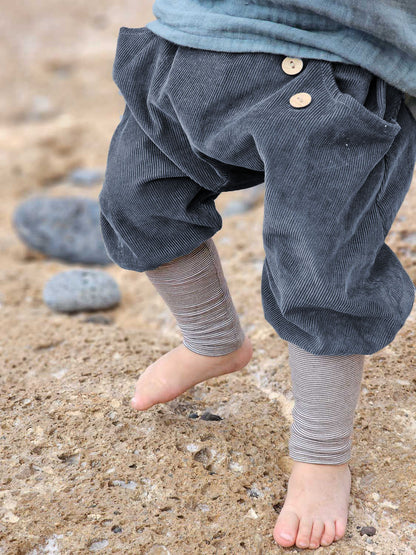 Ein Kind steht barfuß auf Sand, trägt Knickerbocker Feincord Ringel - Blaubeere, sichtbar sind die locker sitzenden Cordhosen mit Holzknöpfen und gestreiften Leggings darunter, ideal für aktive Entdecker:innen.