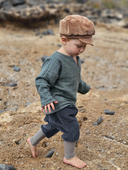 Kleinkind am Strand in Knickerbocker Feincord Ringel - Blaubeere, mit gestreiftem Bund und lockerer Passform, kombiniert mit einem grünen Hemd, entdeckt barfuß die steinige Uferlinie.