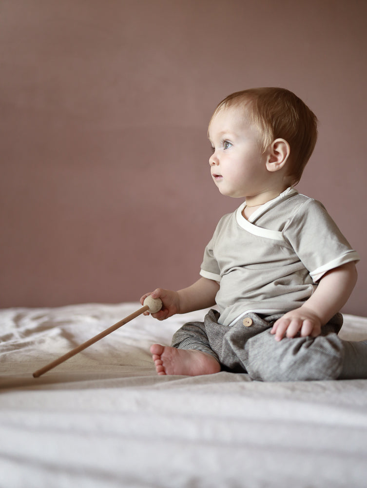 Ein Baby sitzt auf einem Bett und hält einen Holzstab, während es ein kurzes Wickelshirt aus Bambusviskose trägt. Perfekt für Sommer, antibakteriell und handgenäht in Berlin.