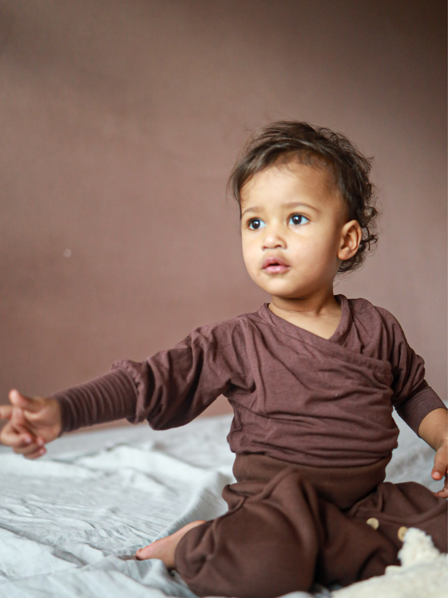 Kleinkind in Knickerbocker Bio-Sweat - Schoko, sitzt auf weichem Untergrund. Die braune, langärmelige Kleidung zeigt den bequemen, legeren Schnitt der Hose, perfekt für aktive Entdecker:innen.