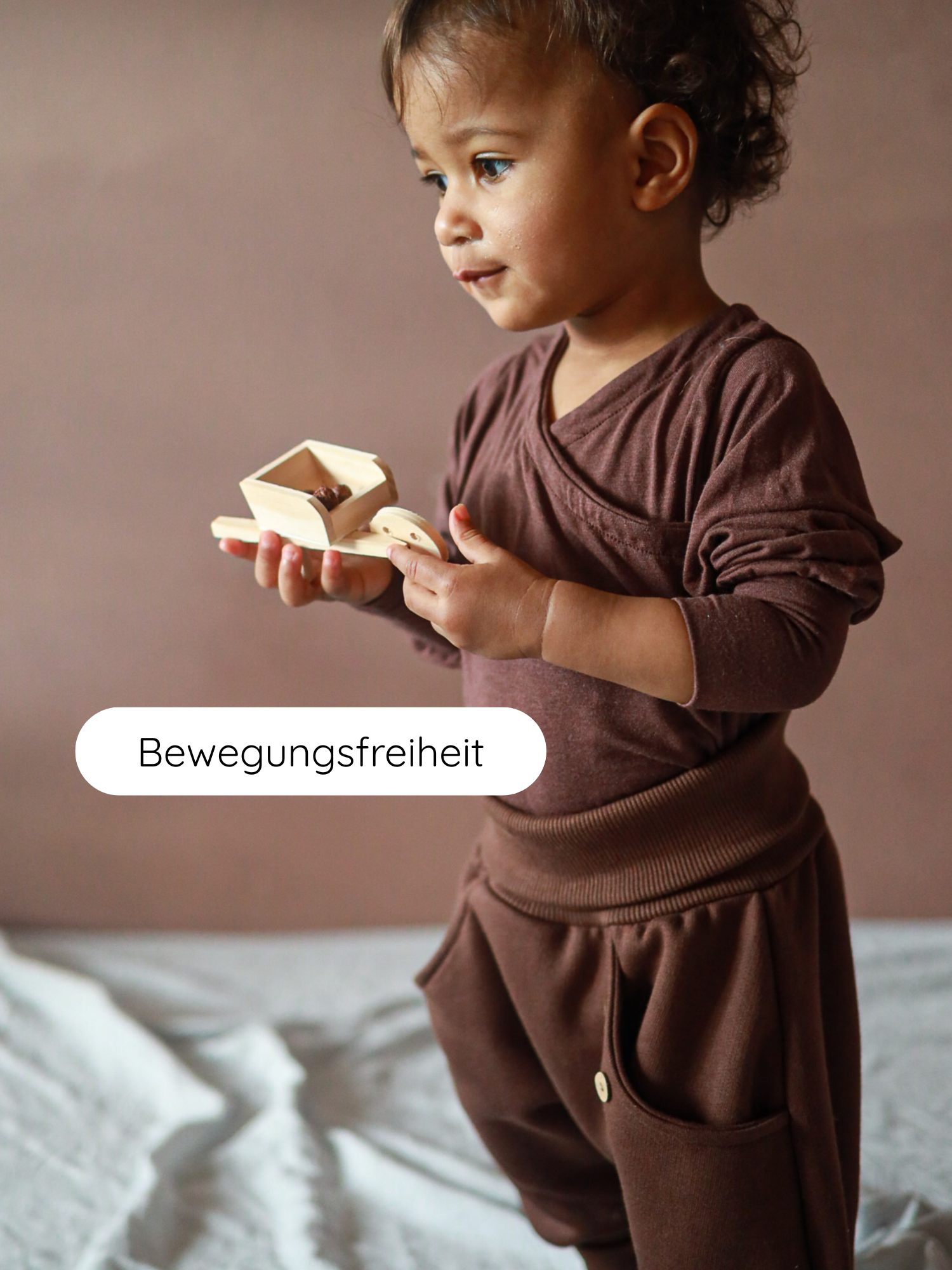 Kleinkind in warmer Knickerbocker Bio-Sweat - Schoko steht auf weichem Untergrund, hält einen kleinen Holzspielzeugkarren. Das Bild vermittelt Bewegungsfreiheit und Gemütlichkeit für kleine Entdecker.
