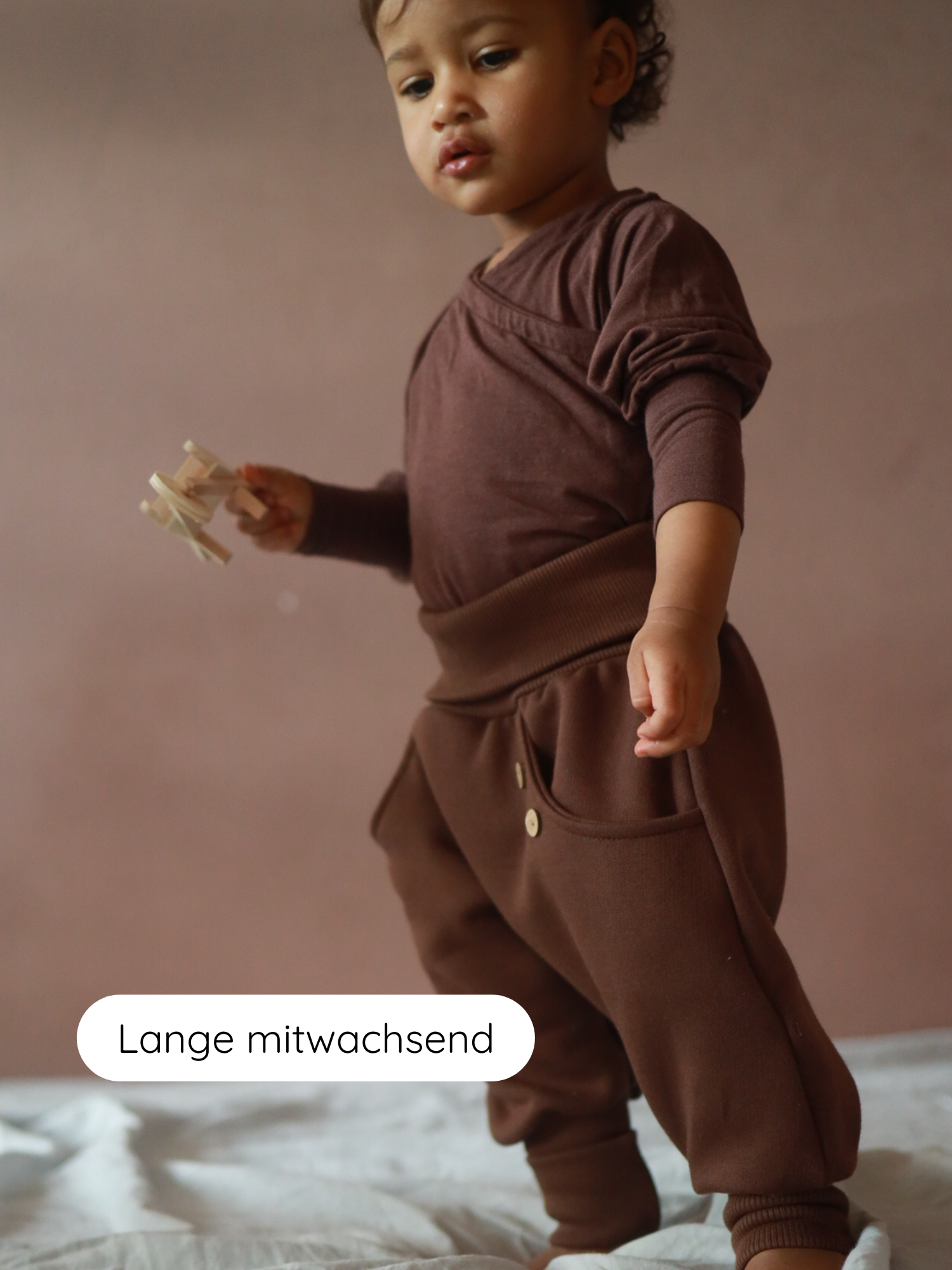 Ein Kleinkind trägt die Knickerbocker Bio-Sweat - Schoko, bestehend aus einem braunen Langarmshirt und lockerer Hose. Es hält ein kleines Holzspielzeug, während es auf einer weichen Oberfläche steht.