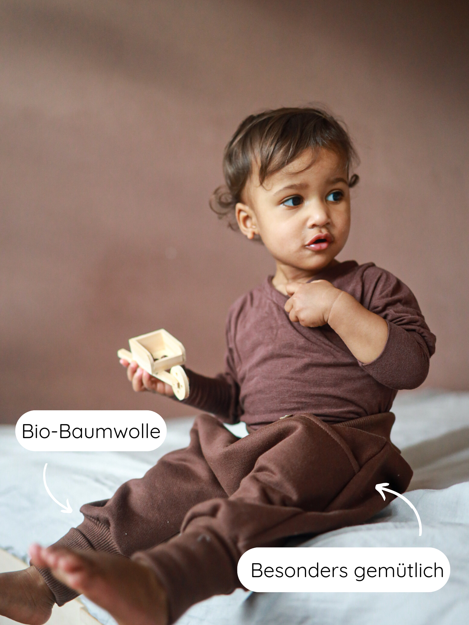 Ein Kleinkind sitzt in einem Schoko-Knickerbocker Bio-Sweat und hält ein kleines Holzspielzeug. Bio-Baumwolle und Besonders gemütlich heben die nachhaltigen und bequemen Eigenschaften des Outfits hervor.