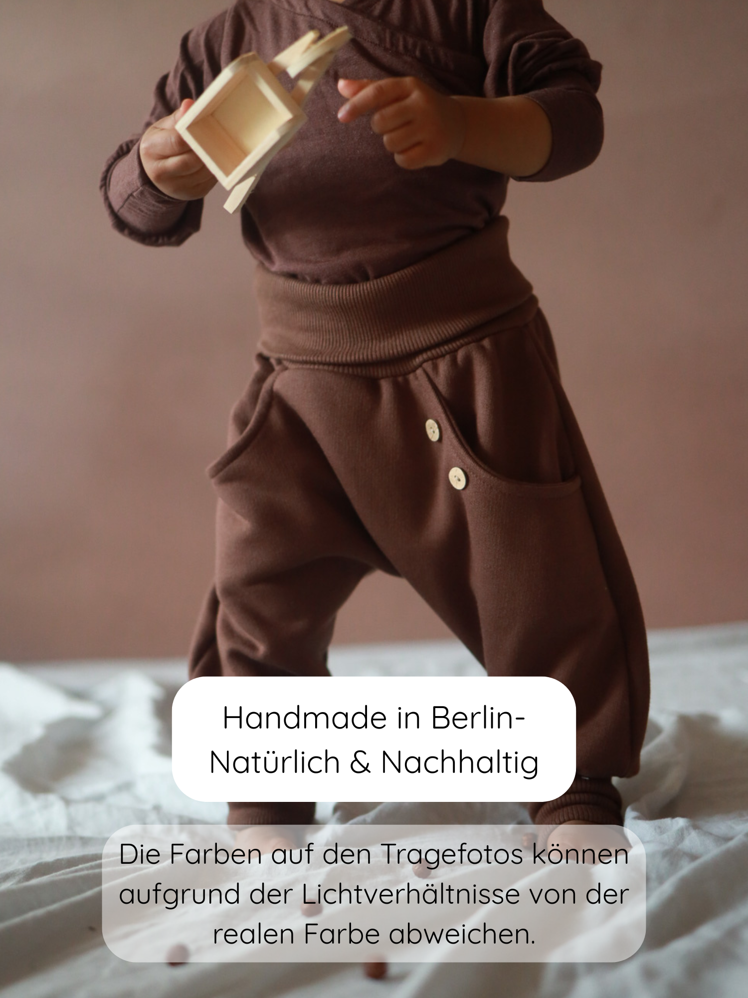 Ein Kind trägt den Knickerbocker Bio-Sweat - Schoko, ein locker sitzendes, zweiteiliges Outfit aus weicher Bio-Baumwolle, und hält ein kleines Holzspielzeug, handgemacht in Berlin.