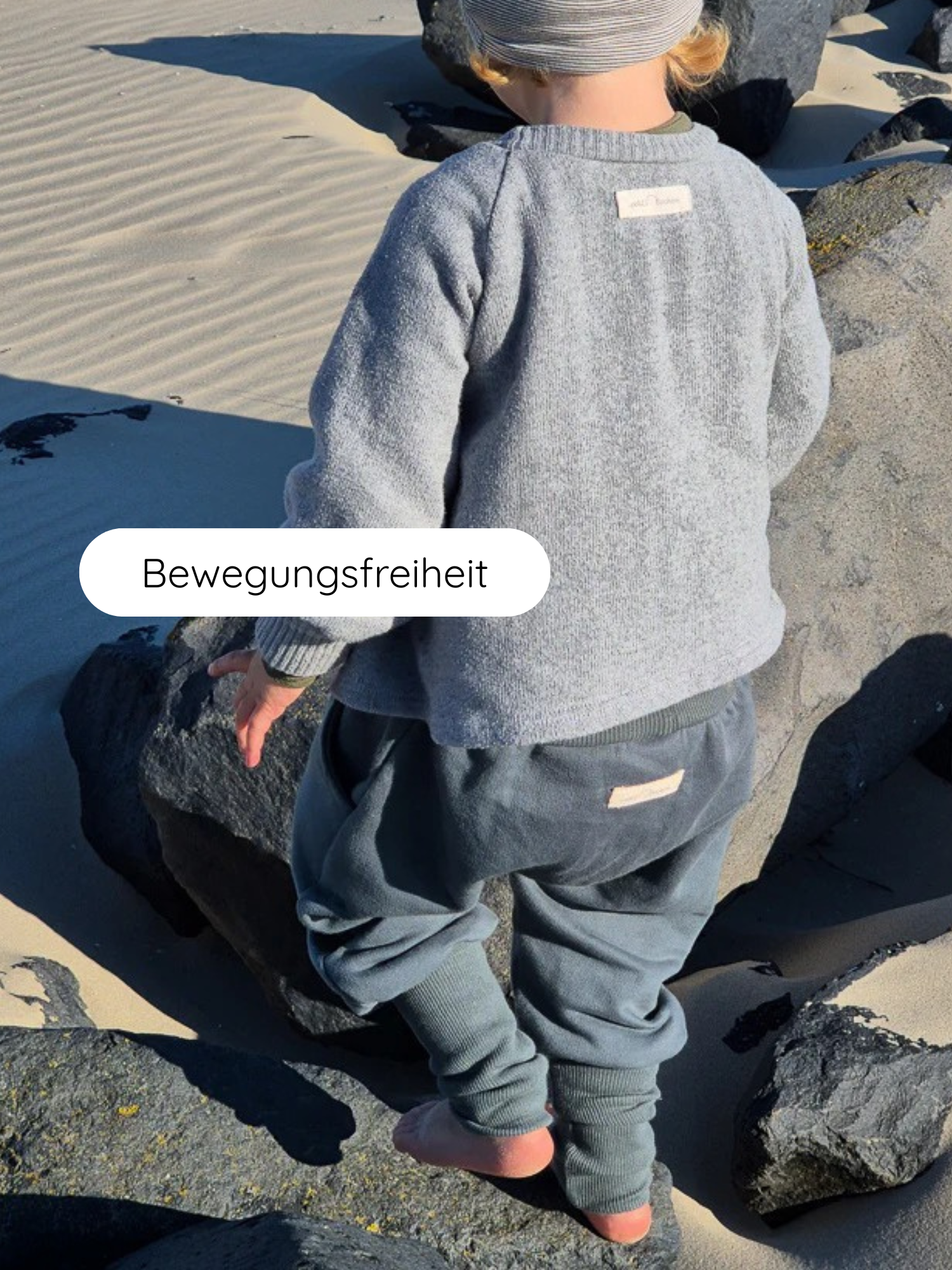Ein Kind in Knickerbocker Bio-Sweat - Taube überquert barfuß dunkle Felsen am Strand, verkörpert Bewegungsfreiheit und Bequemlichkeit durch den elastischen Bund und weichen, atmungsaktiven Stoff.